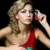 Portrait of a young woman with blonde hair wearing a red top, looking at the camera with her hand near her ear.